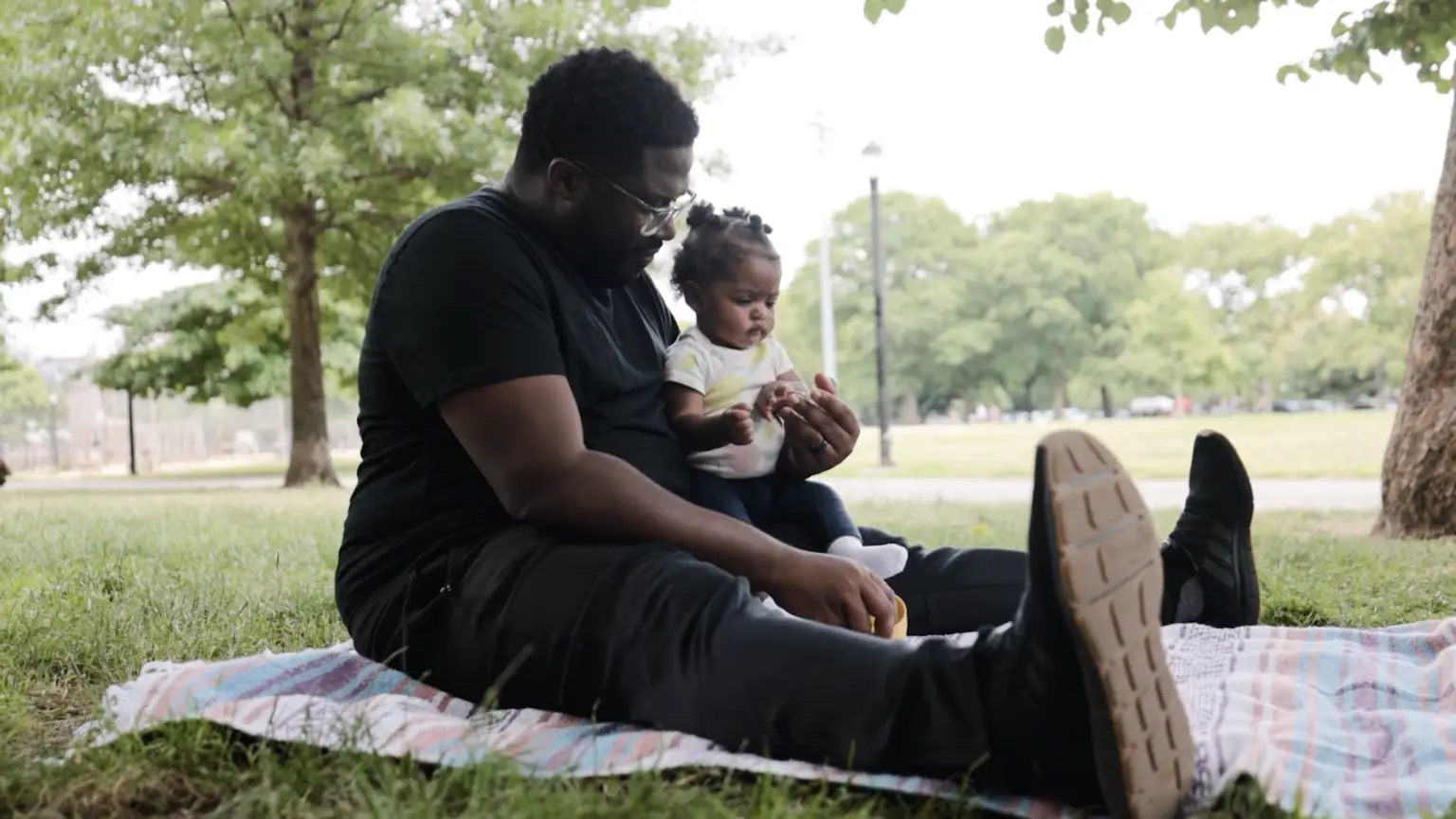 A father and child share a peaceful moment on a blanket in the park, surrounded by green grass and trees.